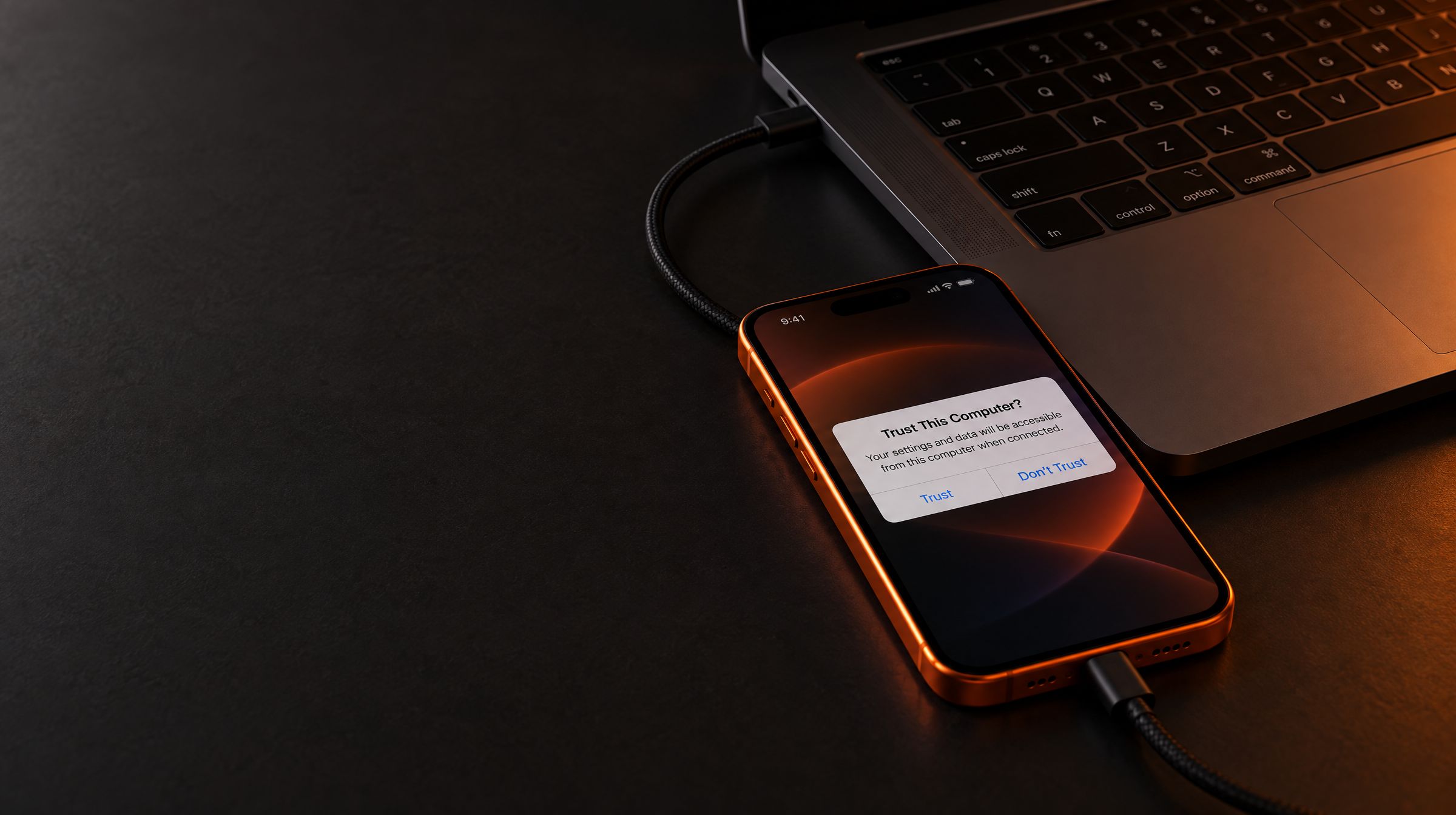 An orange iPhone plugged into a MacBook with a braided USB-C cable, showing the Trust This Computer? prompt.